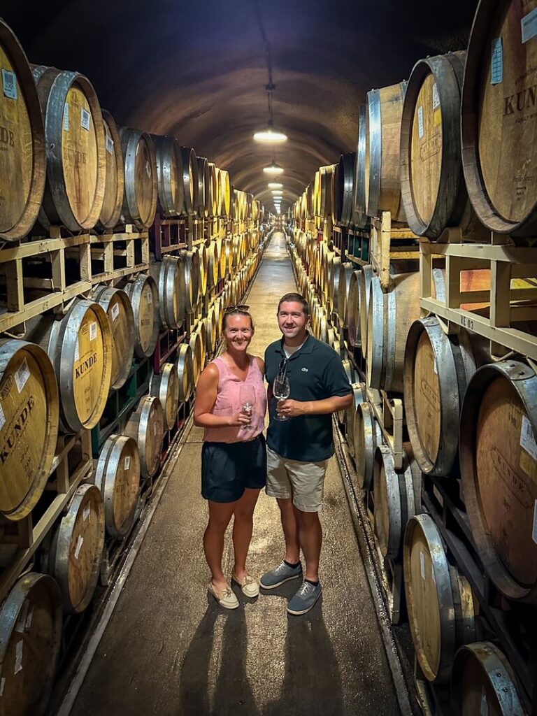 Two visitors stand in the Kunde Family Winery Cave surrounded by wine barrels.