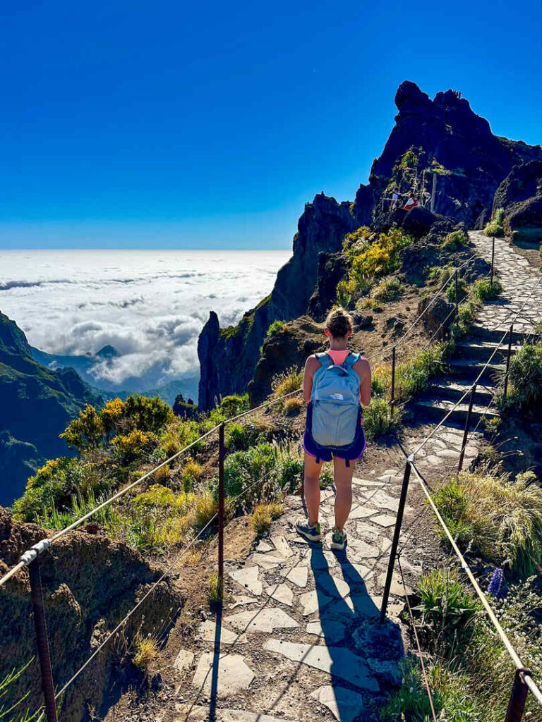 A hiker standing on the trail above the clouds with a mountain peak in the distance under blue skies.