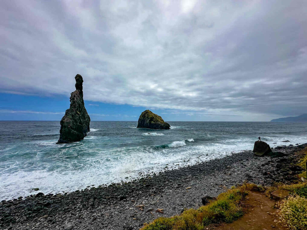 A view of Ilhéus da Ribeira da Janela from the beach. A person stands on a nearby rock, showing the massive scale.