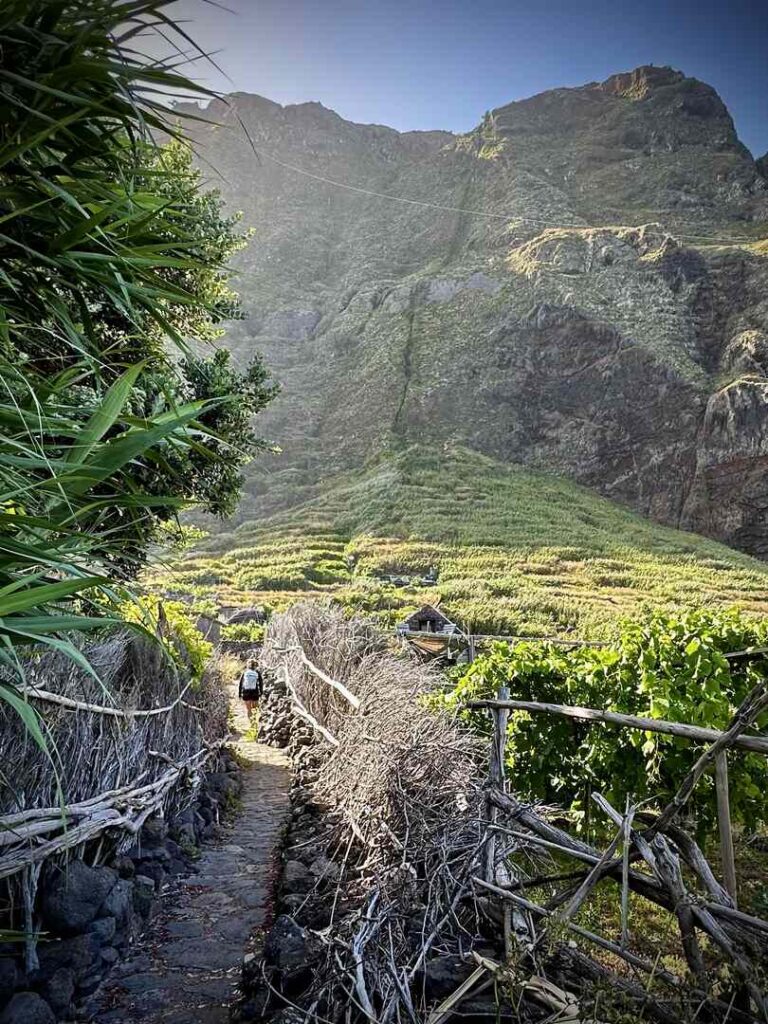 A hiker walking up a narrow path with a large cliff in the background to explore the village.